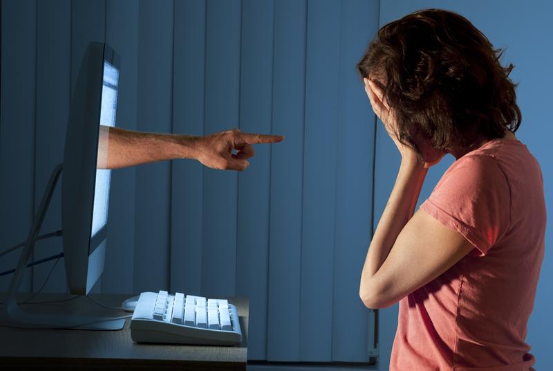 Severely distraught young woman sitting in front of a computer with a judgemental hand pointing at her from within the computer monitor being cyber bullied.